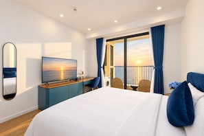 A hotel room with a large bed, a TV, a desk, and a balcony with a view of the sunset. - Skywave Apartment Firework Delight (Phu Quoc)