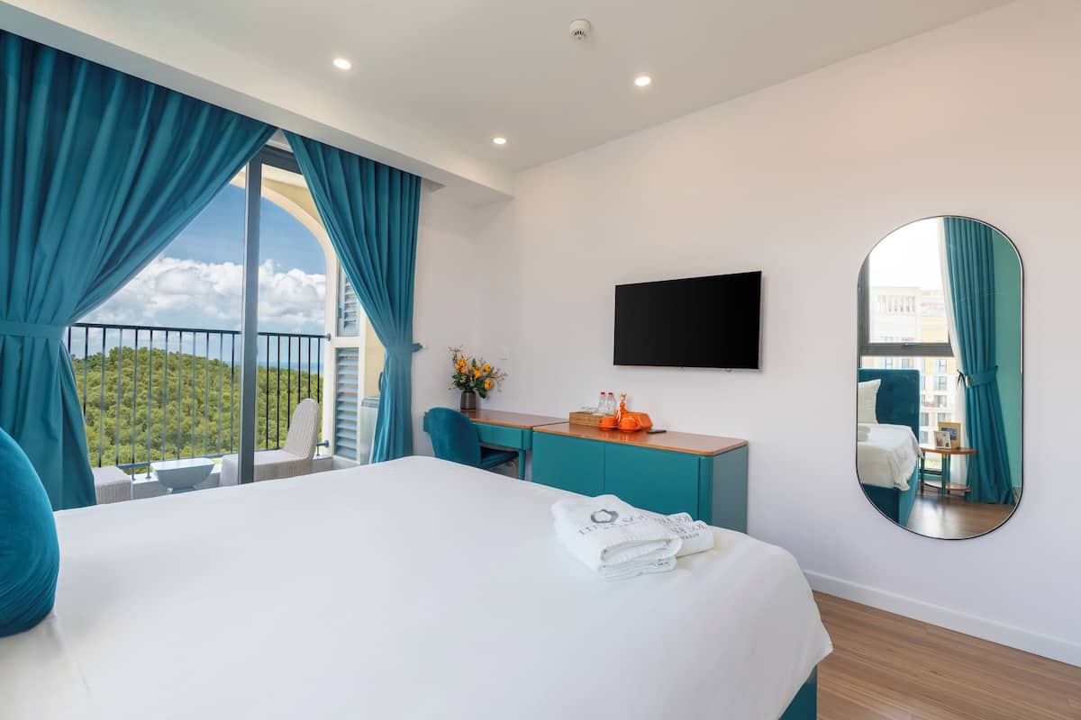 Une chambre d’hôtel avec un grand lit, un bureau, une chaise, une télévision et un balcon avec des rideaux bleus.