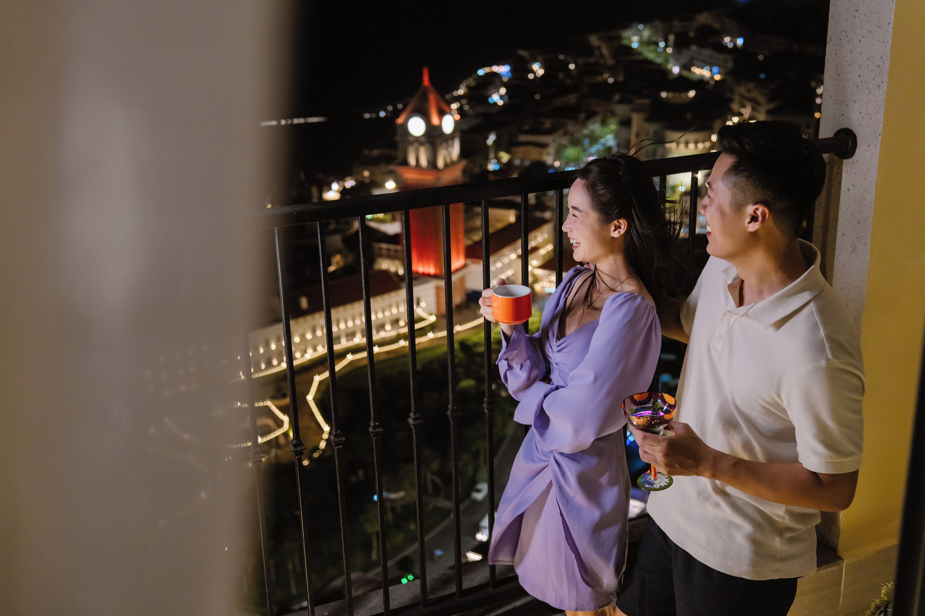 A couple standing on a balcony overlooking a cityscape with a bridge and a tower.