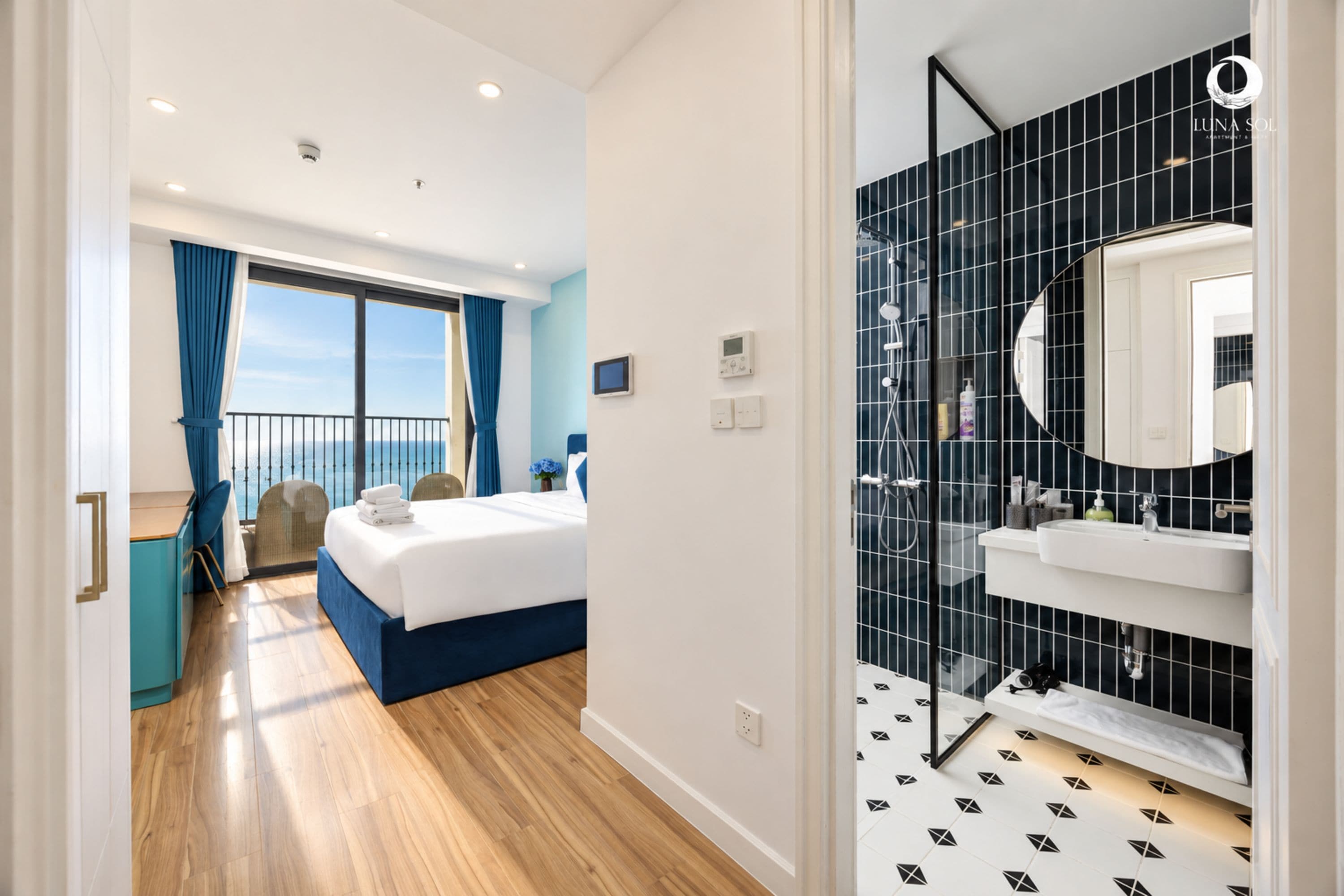A hotel room with a bed, a balcony with curtains, a bathroom with a tiled wall, and a view of the sea.