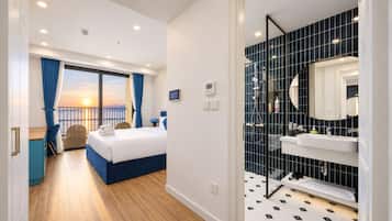 Quarto de hotel com cama, escrivaninha, cadeira, varanda com cortinas e banheiro com espelho e pia.