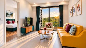 A modern living room with a sofa, a coffee table, and a television. There is a balcony with a view of greenery.