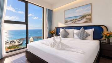 Quarto de hotel com uma janela grande que oferece vista para o mar e um cais, uma cama bem arrumada com toalha, e um quadro com cena de praia.