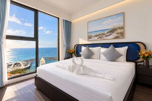 Habitación de hotel con un ventanal que ofrece vistas al mar y un muelle, una cama bien tendida con una toalla, y un cuadro con una escena de playa.