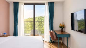Habitación de hotel con cama, escritorio, silla, televisión y balcón con vista a árboles.