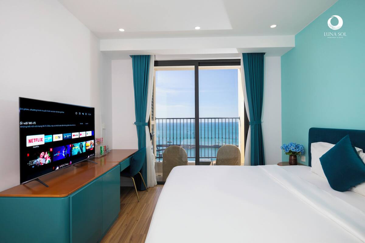 Une chambre d’hôtel moderne dotée d’une grande télévision diffusant des services de streaming, d’un balcon avec vue sur la mer et d’un lit soigneusement fait avec des oreillers bleus.