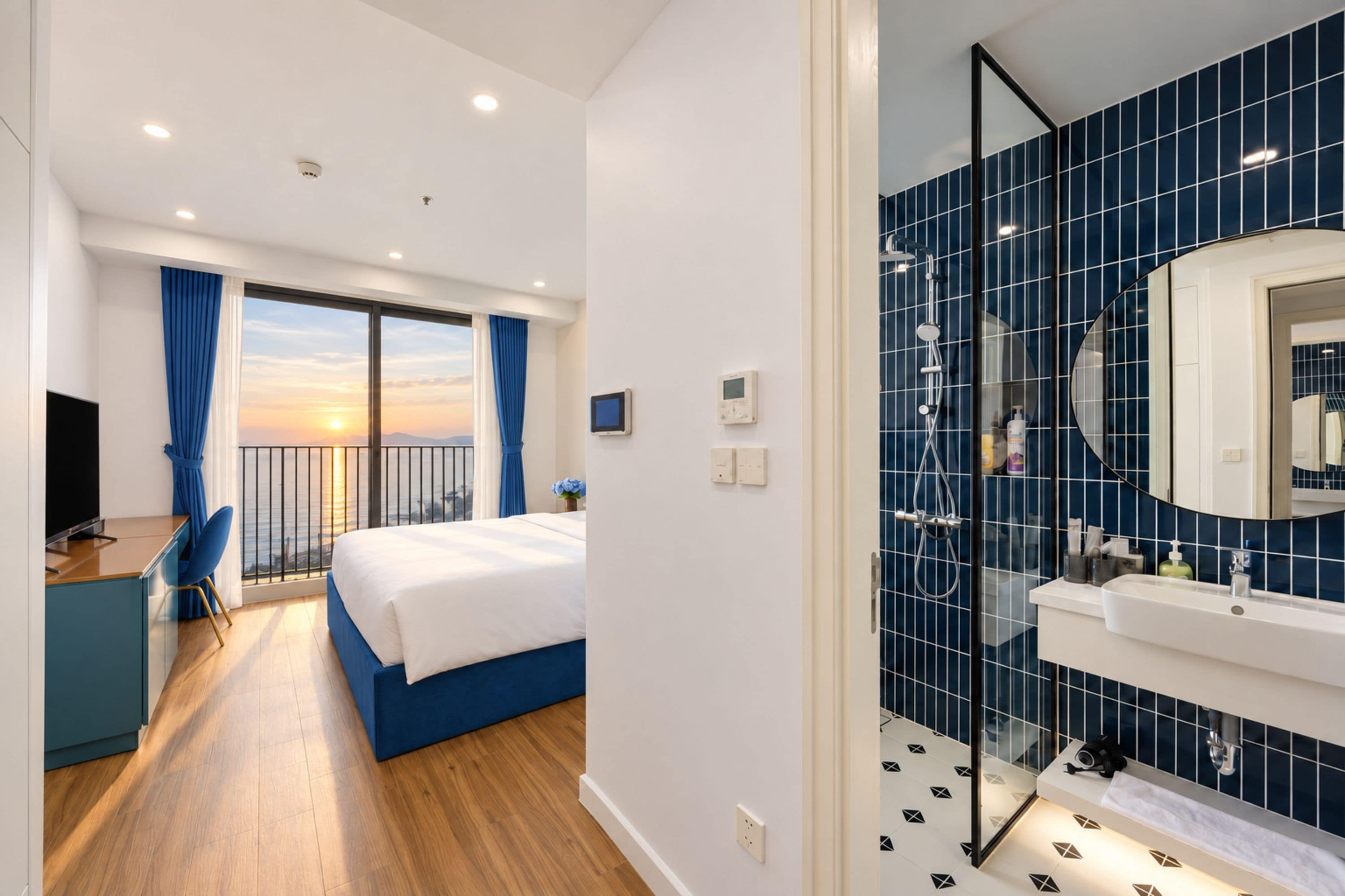 A hotel room with a balcony, a bed, a desk, and a bathroom with a large mirror and blue tiles.