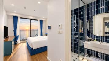 A hotel room with a balcony, a bed, a desk, and a bathroom with a large mirror and blue tiles.