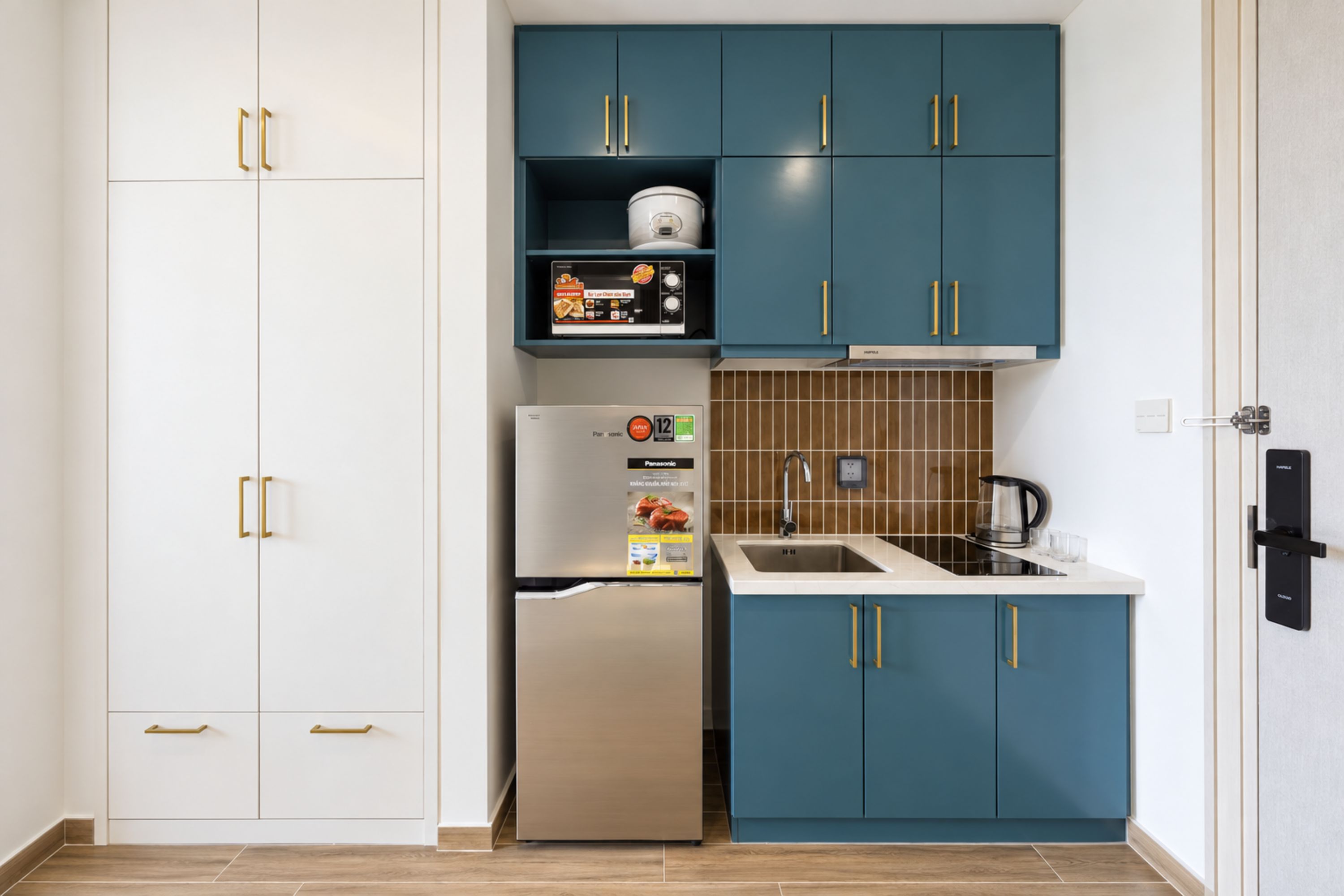 Une cuisine compacte avec des meubles blancs et turquoise, un réfrigérateur en acier inoxydable et une bouilloire noire posée sur le plan de travail.