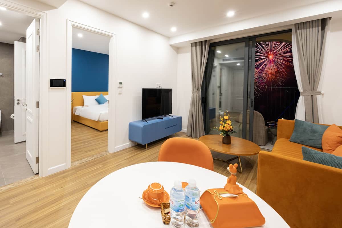 Une chambre d’hôtel avec un plancher en bois, un lit avec une literie bleue, une télévision, un canapé, une table avec un vase et un jouet, et une vue sur les feux d’artifice.