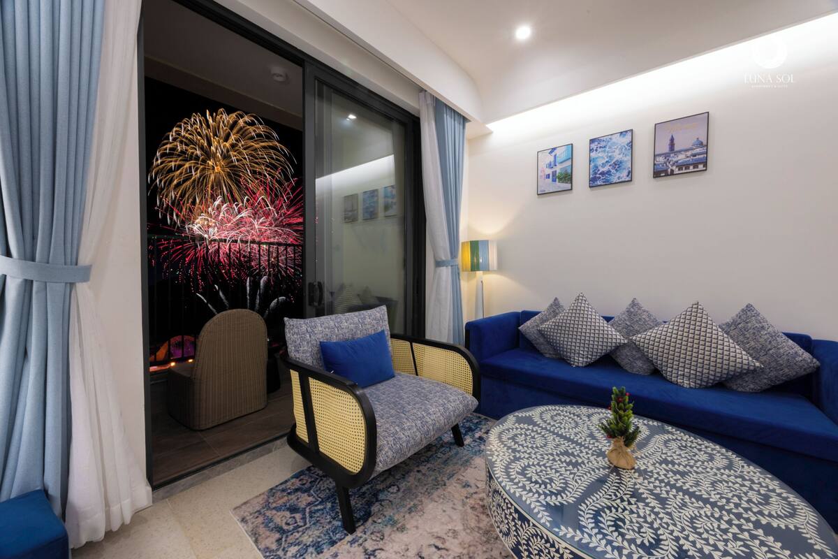 Un salon avec un canapé, un fauteuil et une table basse, un petit sapin de Noël et une grande fenêtre donnant sur un spectacle de feux d’artifice.