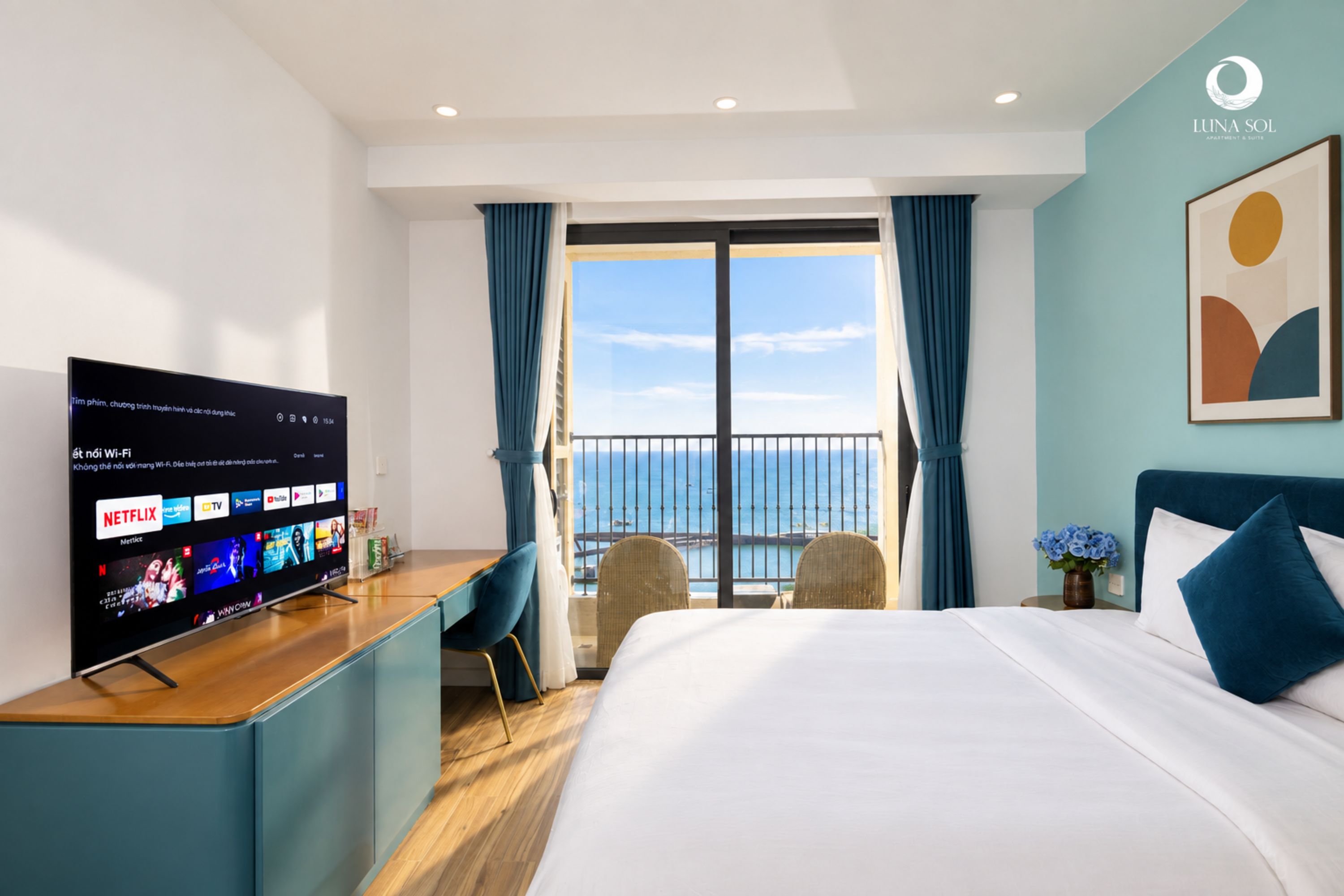 Une chambre d’hôtel moderne dotée d’un grand lit, d’un téléviseur à écran plat permettant d’accéder aux services de diffusion en continu, d’un balcon avec vue sur la mer et d’une décoration aux tons bleus.