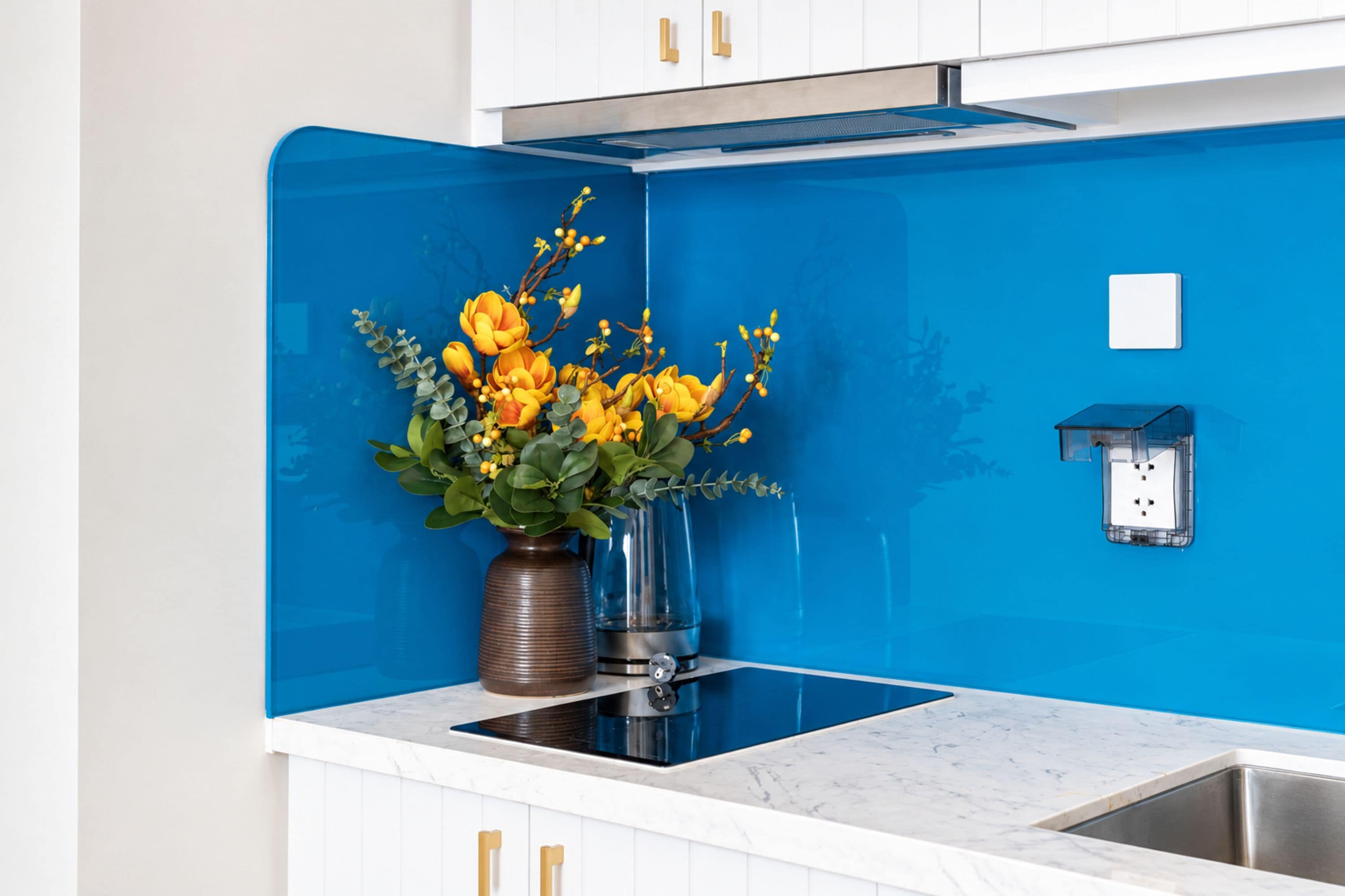 A kitchen with a blue backsplash, a marble countertop, a stainless steel sink, and a vase with yellow flowers.