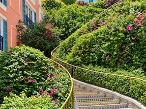 A curved staircase with patterned tiles, surrounded by lush greenery and pink flowers, leading up to a building with blue shutters. - Skywave Apartment Firework Delight (Phu Quoc)