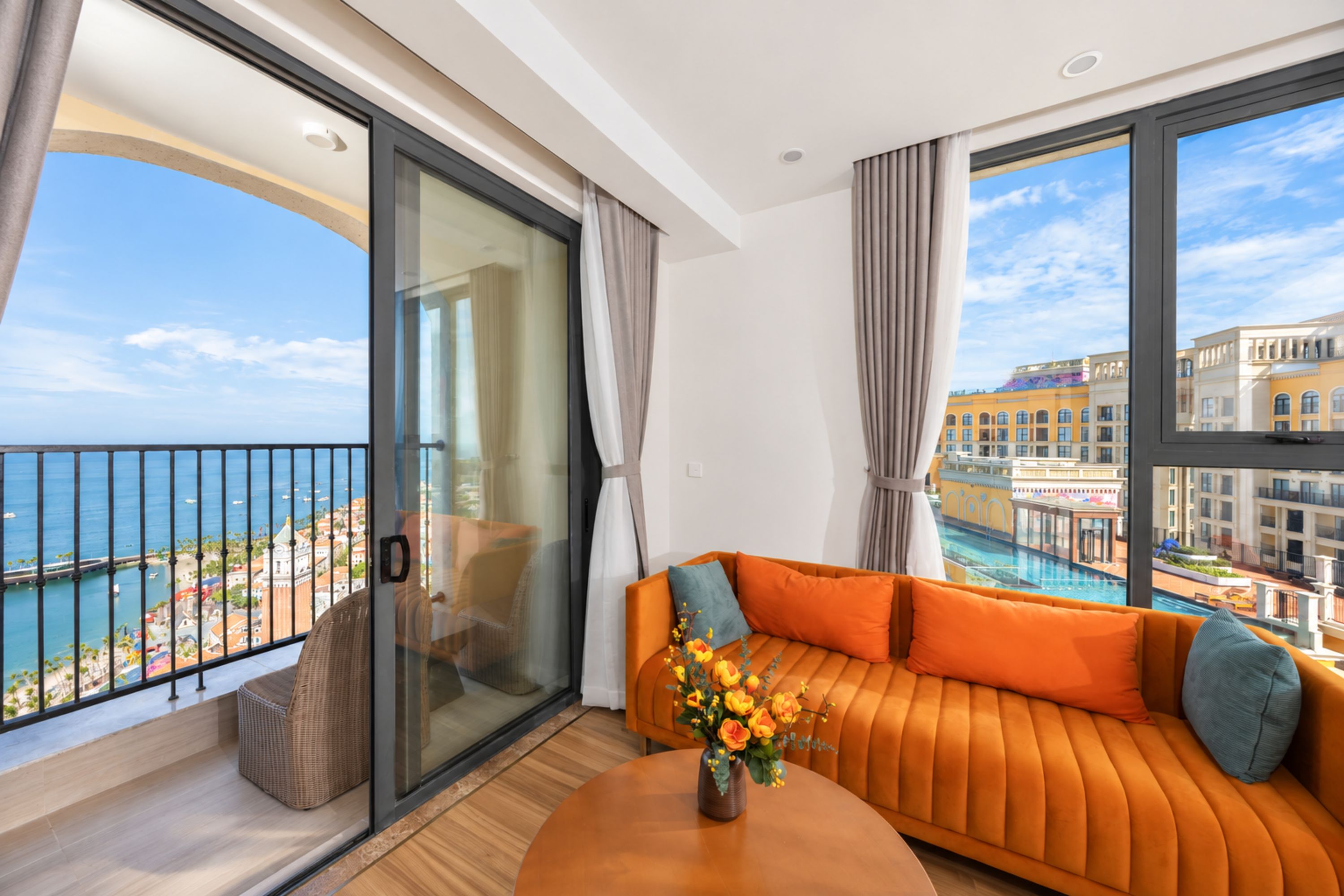 A modern hotel room with a balcony, a sofa, and a view of the sea and buildings.