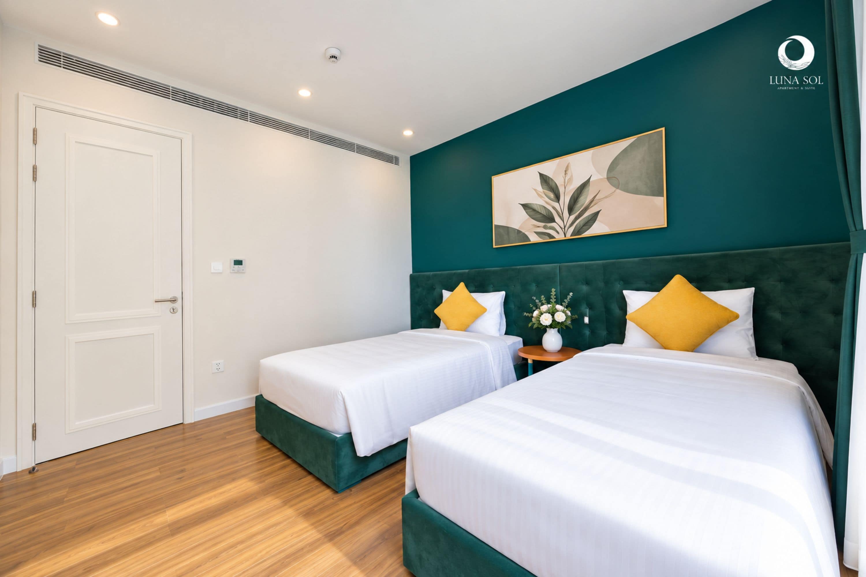 Una habitación de hotel con dos camas, una pared de acento verde, una obra de arte enmarcada y una puerta blanca.