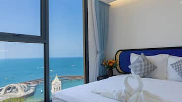 A hotel room with a large window offering a view of the sea and a bridge. The bed has white linens and decorative pillows.