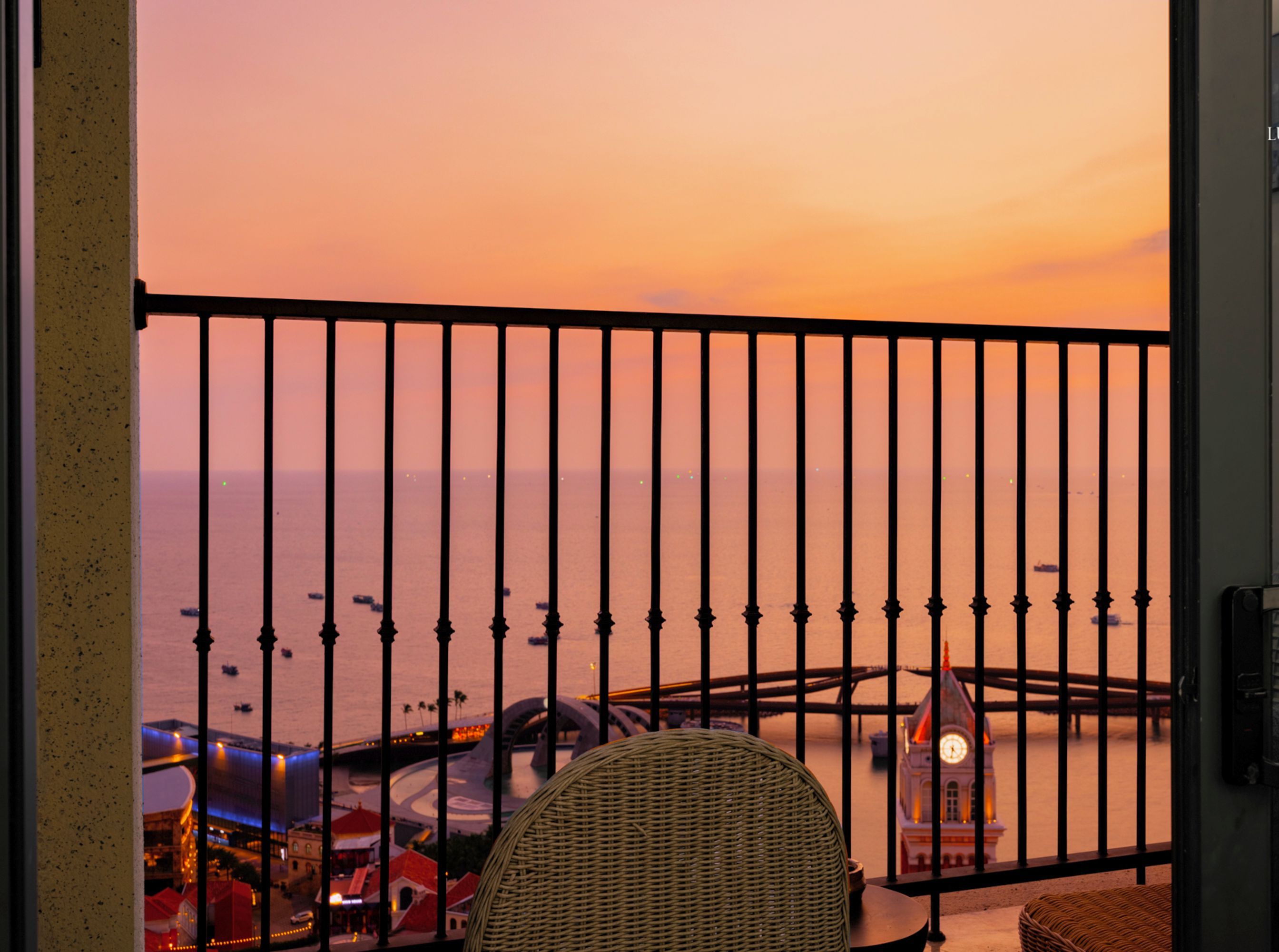 A balcony with a view of the sea at sunset, a wicker chair, and a table with a clock.
