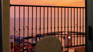 Un balcon avec vue sur la mer au coucher du soleil, une chaise en osier et une table avec une horloge.