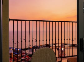Un balcón con vista al mar al atardecer, una silla de mimbre y una mesa con un reloj.
