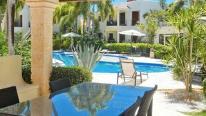 Un patio al aire libre con una mesa y sillas de vidrio, un ventilador de techo y vistas a una piscina y palmeras.