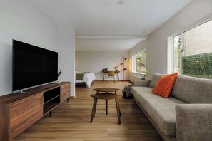 Uma sala de estar moderna com televisão de tela plana, um rack de entretenimento de madeira, um sofá cinza com almofada laranja, uma pequena mesa de madeira e uma grande janela que proporciona iluminação natural.