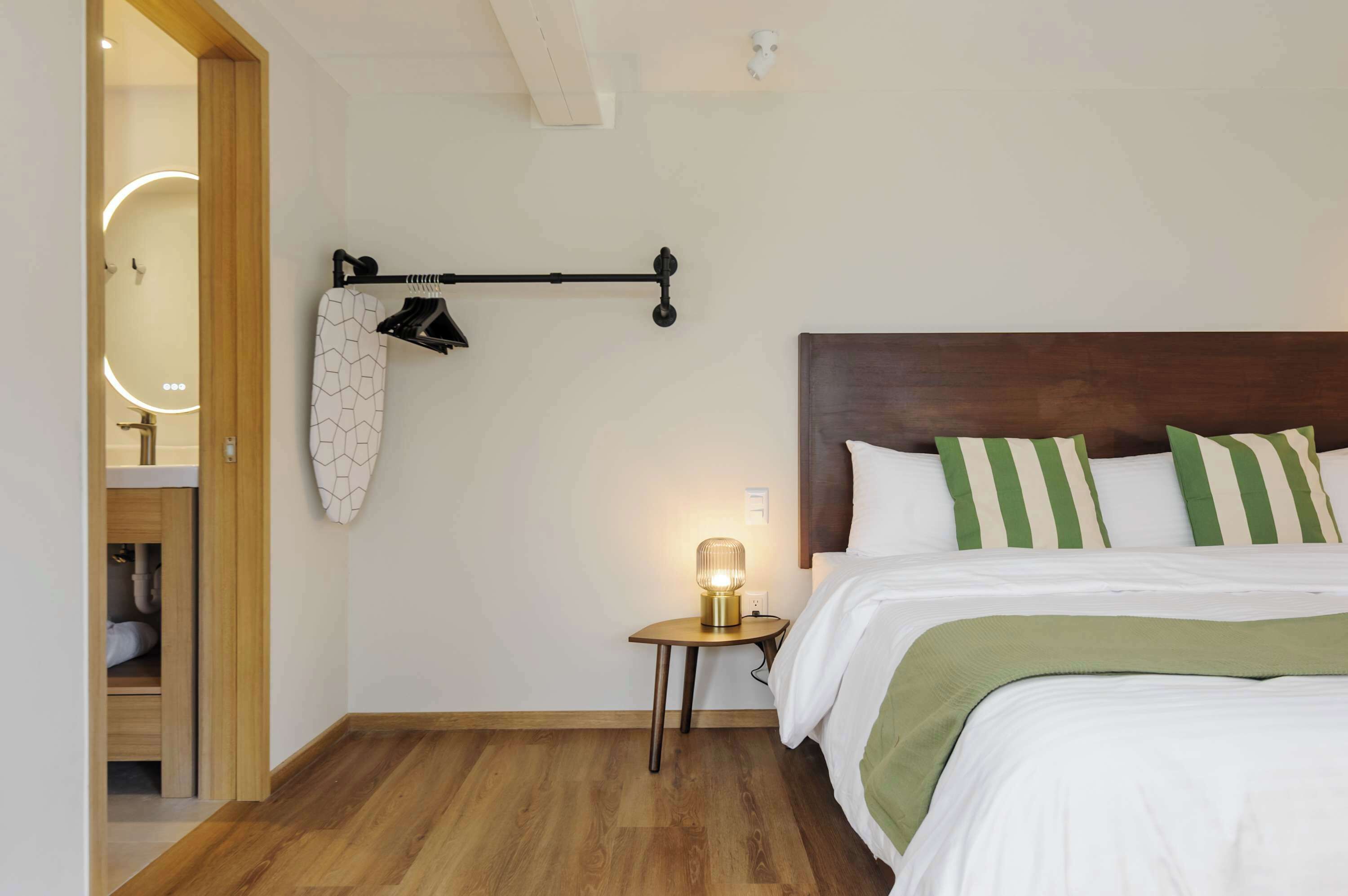 A bedroom with a wooden floor, a bed with green and white striped pillows, a bedside lamp, and a coat rack with a patterned garment.