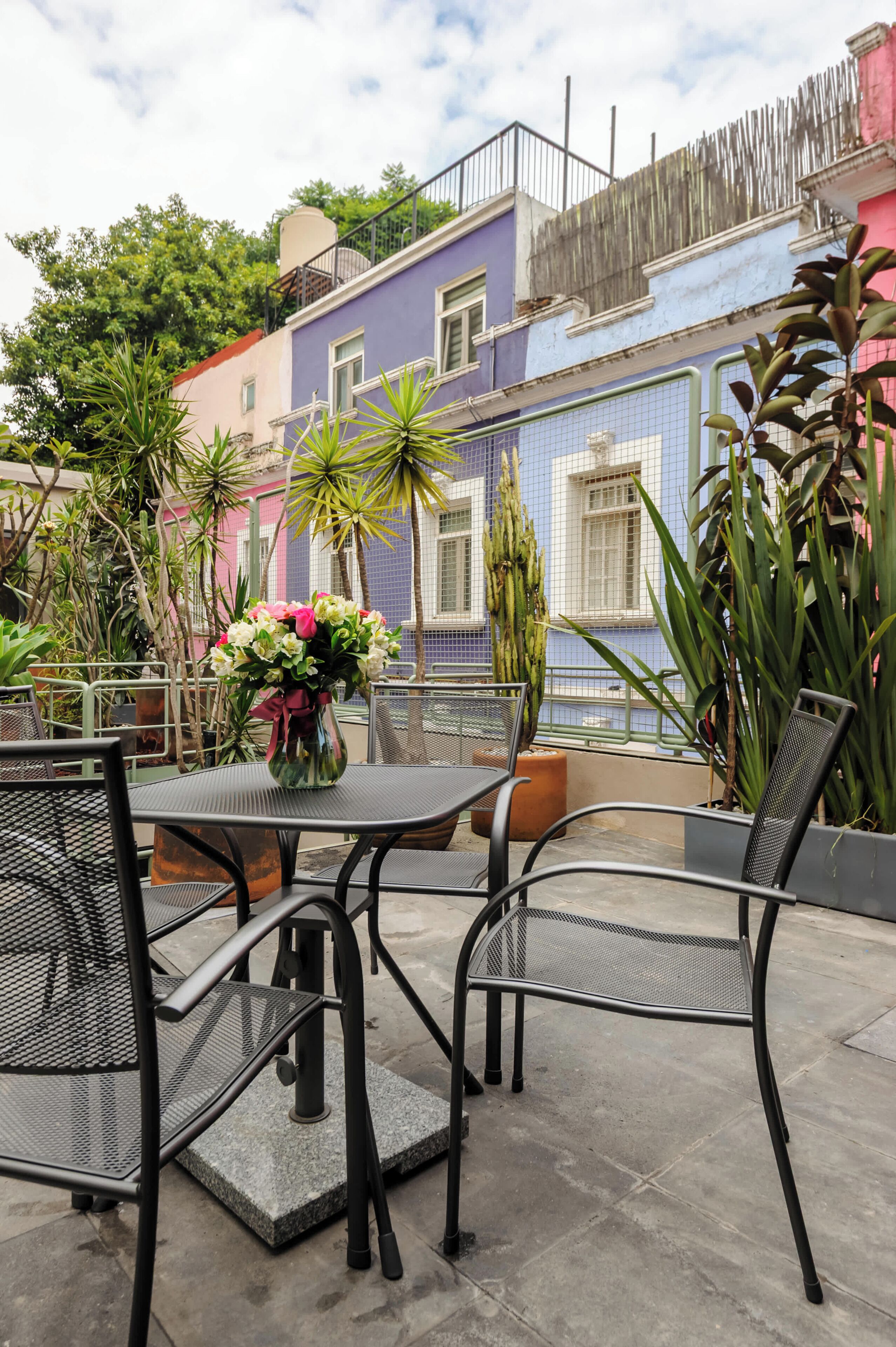 En udendørs patio med metalstole og et bord, omgivet af plantekasser og en farverig bygning i baggrunden.