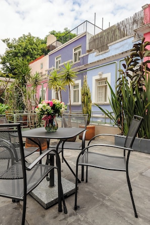 An outdoor patio area with metal chairs and a table, surrounded by potted plants and a colorful building in the background.
