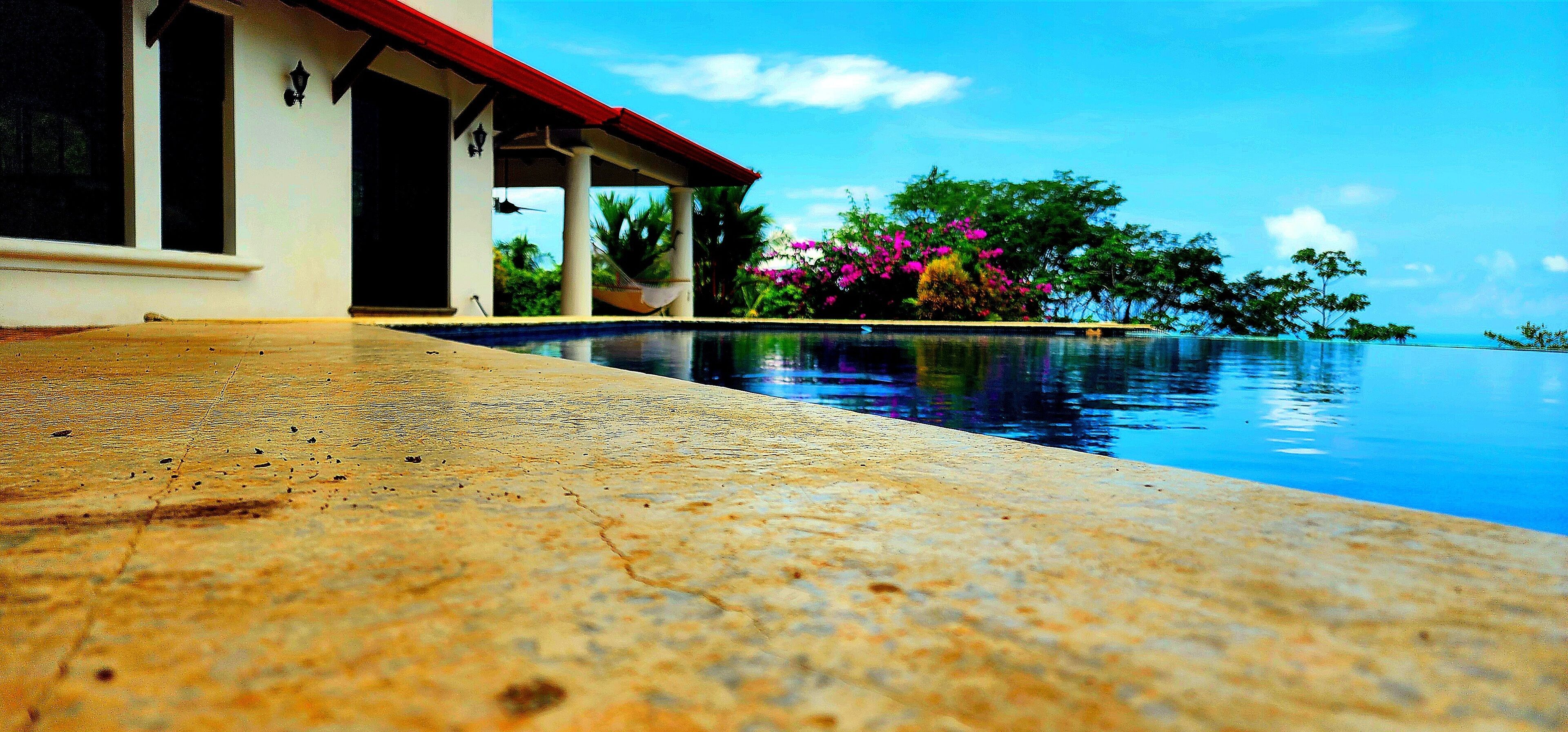 Uma área à beira da piscina com um deck de concreto, um espaço de estar coberto com telhado vermelho, e vista para o oceano e árvores.