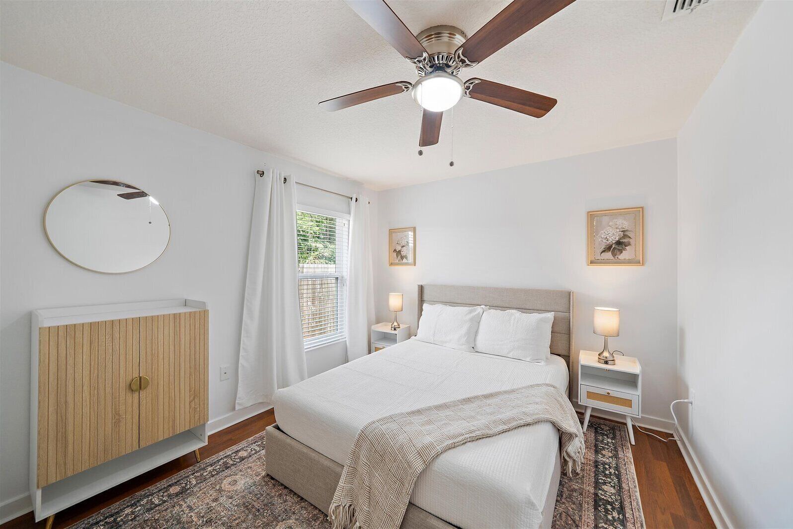 A bedroom with a bed, bedside tables, a ceiling fan, a window with curtains, a round mirror, and a wooden cabinet.
