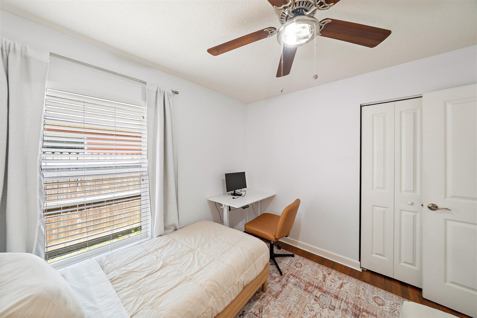 Une chambre à coucher avec un lit, un bureau avec un ordinateur, une chaise, un ventilateur de plafond et une porte.