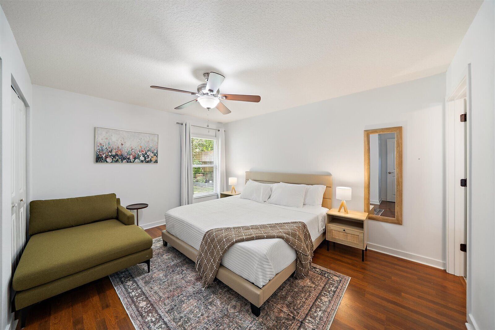 A bedroom with a bed, a chair, a ceiling fan, a nightstand, a lamp, a mirror, and a window with curtains.
