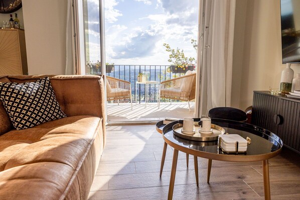A living room with a brown leather sofa, a round coffee table with decorative items, and a balcony with wicker chairs and potted plants.