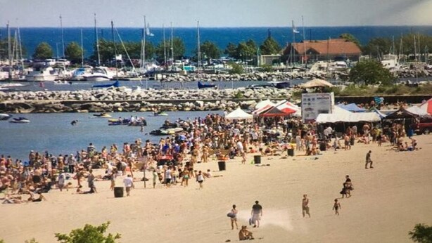 Una playa concurrida, gente y barcos, y un puerto deportivo al fondo.