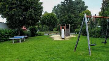 Ein Spielplatz mit Schaukeln, einer Rutsche und einem Tischtennis-Tisch in einer grünen Parklandschaft mit Bäumen.