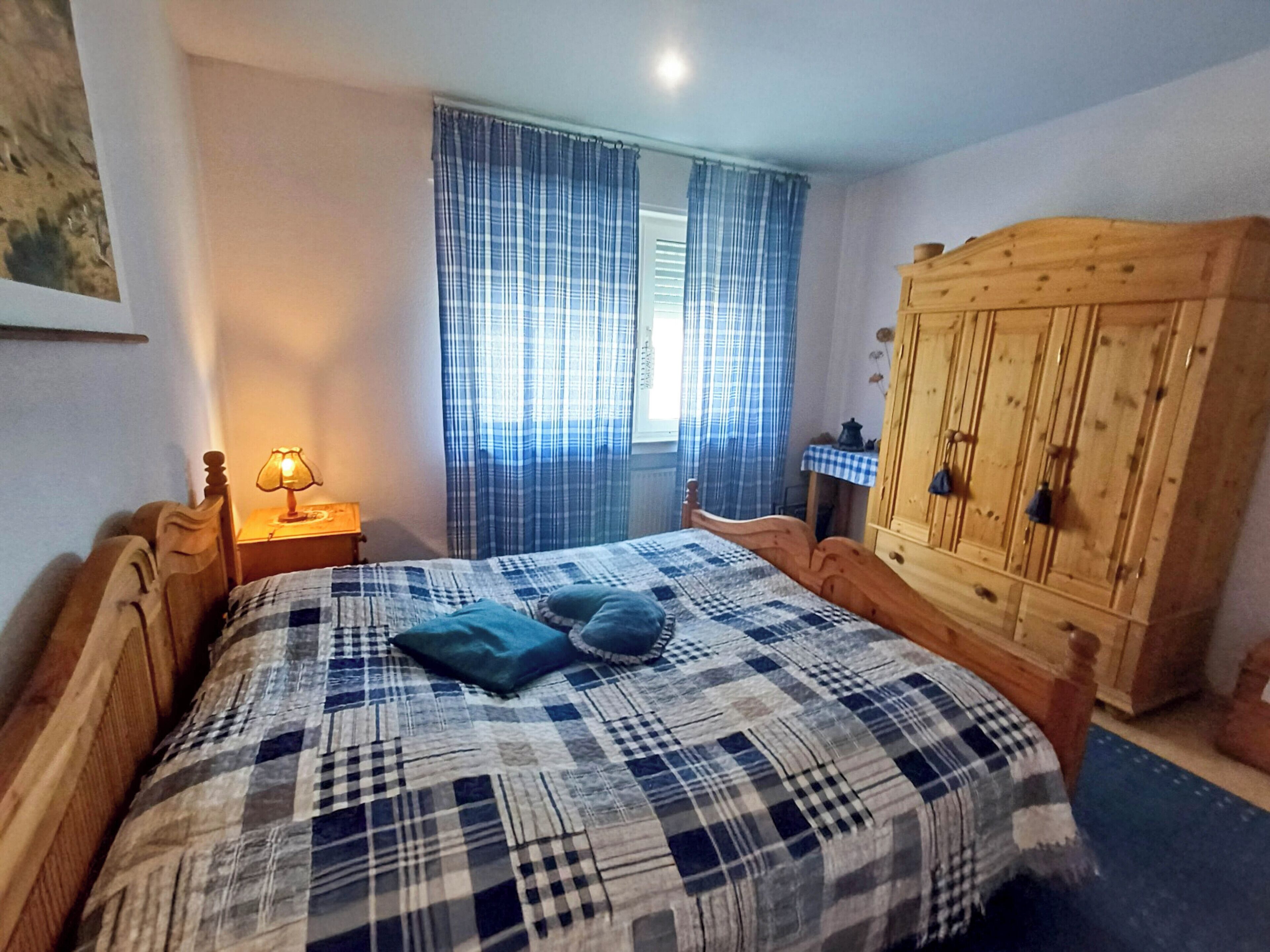 Ein Schlafzimmer mit einem hölzernen Bett, einem Nachttisch mit Lampe, einem Holzschrank und einem Fenster mit Vorhängen.