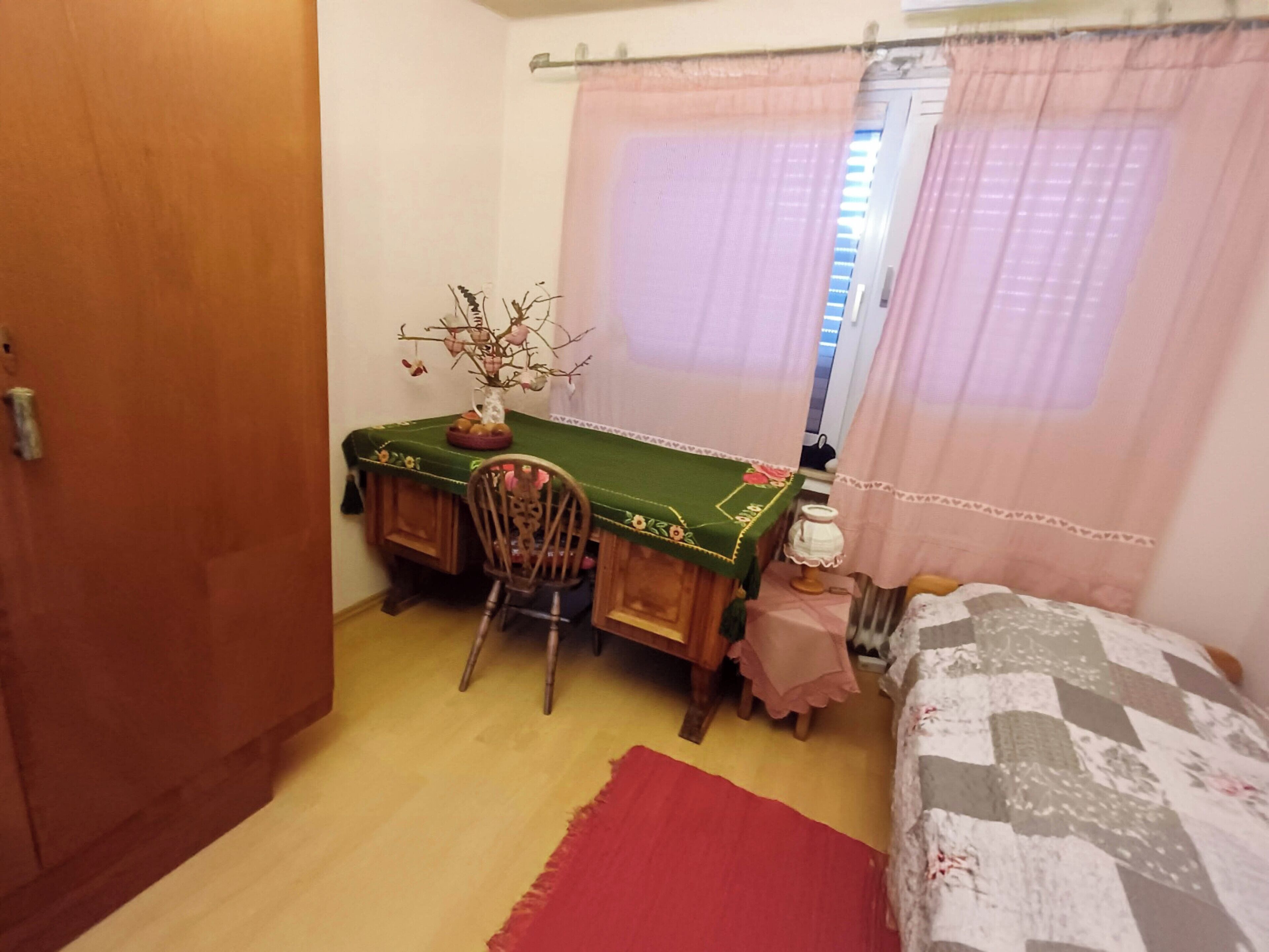 A small bedroom with a wooden desk, a chair, a bed with a quilt, a window with curtains, and a door.