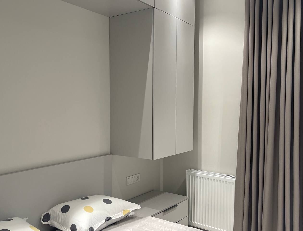 Un dormitorio moderno y minimalista con armario empotrado, radiador y mesita de noche con un cojín de lunares.
