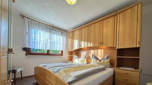 A bedroom with a wooden bed, a nightstand, a wardrobe, and a window with curtains. - Holiday home in the Thuringian Forest (Langenbach)