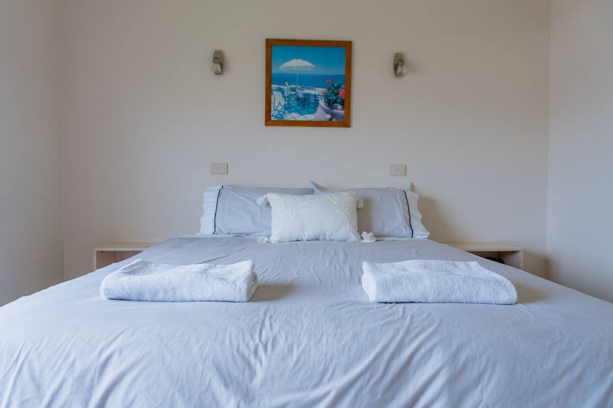 Un lit bien fait, avec du linge de lit blanc et deux serviettes, un tableau représentant un paysage côtier accroché au mur et une simple table de chevet.