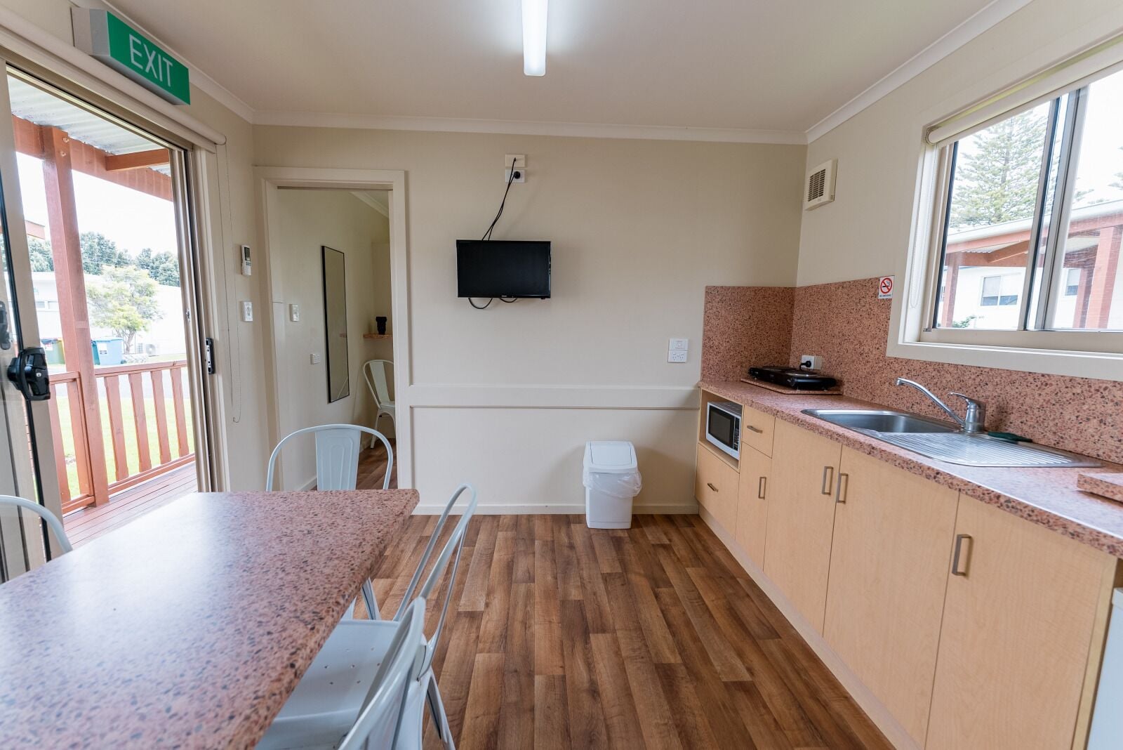 A compact kitchen with wooden flooring, a sink, a microwave, and a television mounted on the wall.