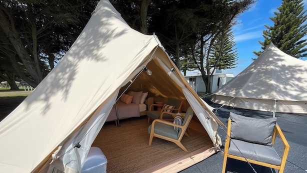 Una tenda con zona notte, comprensiva di un letto e sedie, allestita su una superficie pavimentata, con alberi sullo sfondo.