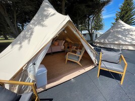 Una tienda de campaña con zona de descanso, que incluye una cama y sillas, montada sobre una superficie pavimentada, con árboles al fondo.