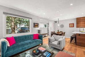 A modern living room with a teal sofa, a grey armchair, and a glass coffee table. There is a dining area with a wooden table and chairs, and a kitchen area with a wooden countertop and sink.