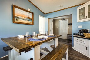 Un comedor con una mesa de madera dispuesta para cuatro personas, una banca y una zona de cocina con armarios blancos y una cafetera.