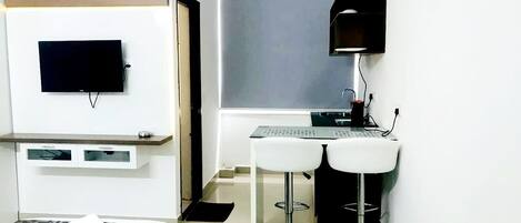A compact hotel room with a bed, a TV mounted on the wall, a desk with a computer, and two white bar stools.