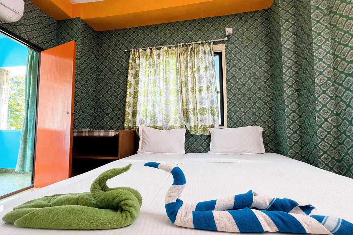 A hotel room with a bed, a green and blue striped towel, a green pillow, and a window with patterned curtains.