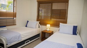 A twin room with two single beds, wooden blinds, a bedside table with a lamp, and a window with blinds.