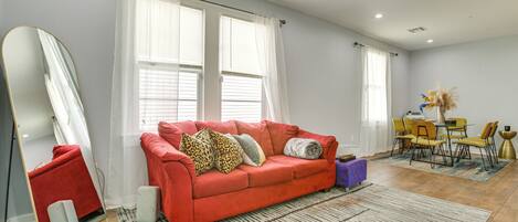 A living room with a red sofa, a large mirror, a dining table with chairs, and a patterned rug.